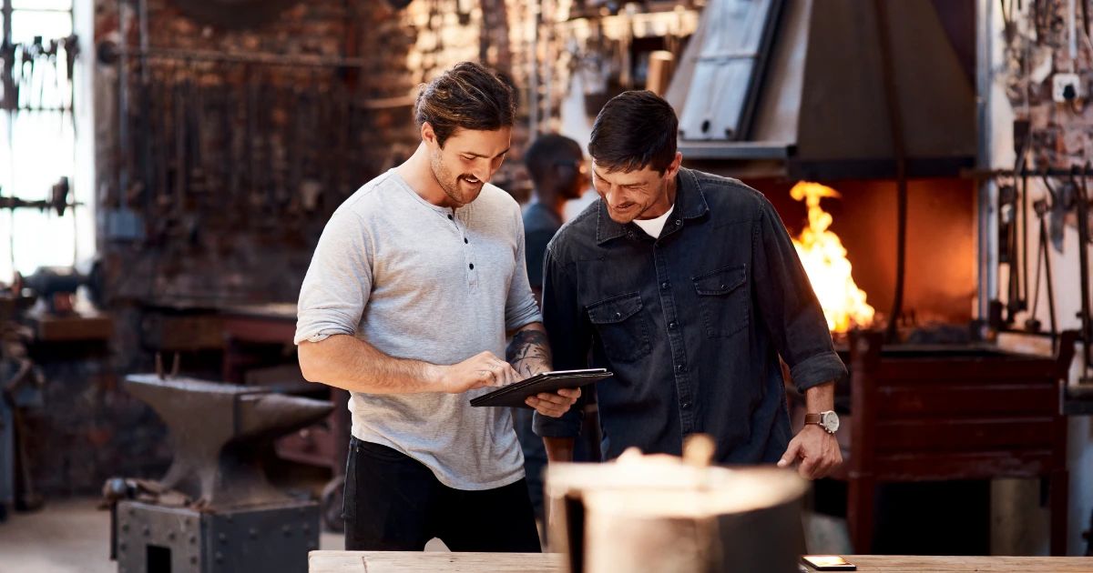 Two men reviewing a tablet together in a working blacksmith forge, anvil and open furnace flames visible behind them