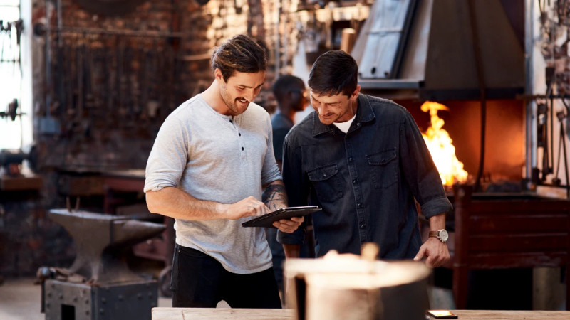 Two men reviewing a tablet together in a working blacksmith forge, anvil and open furnace flames visible behind them
