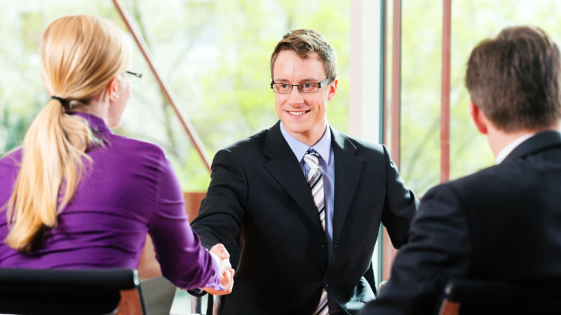 Smiling man in suit and tie shaking hands across a table with two interviewers in a bright office