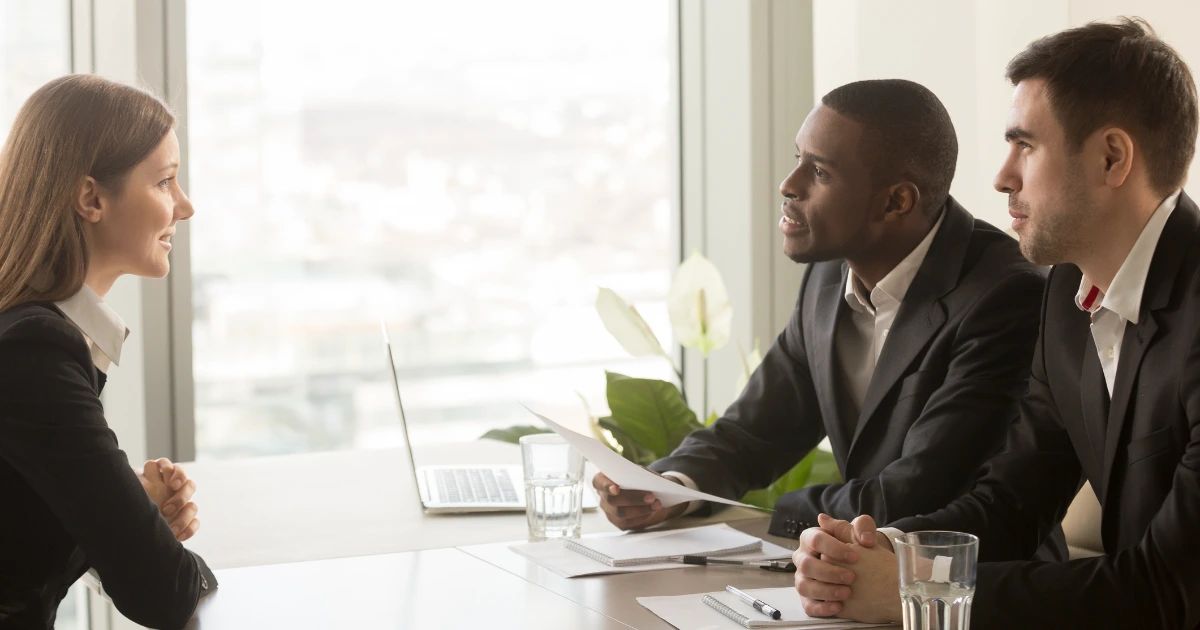 Two suited interviewers at a meeting table speaking with a smiling woman opposite, laptop and papers visible