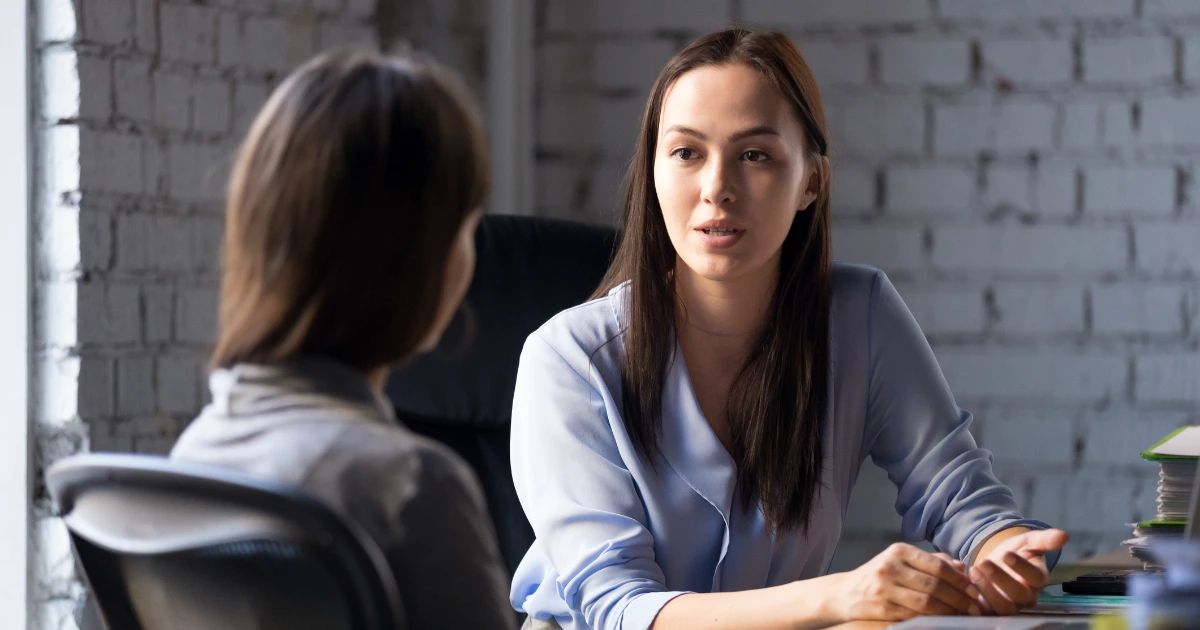 Two women in conversation across a desk in a whitewashed brick office, one facing camera in a light blue shirt