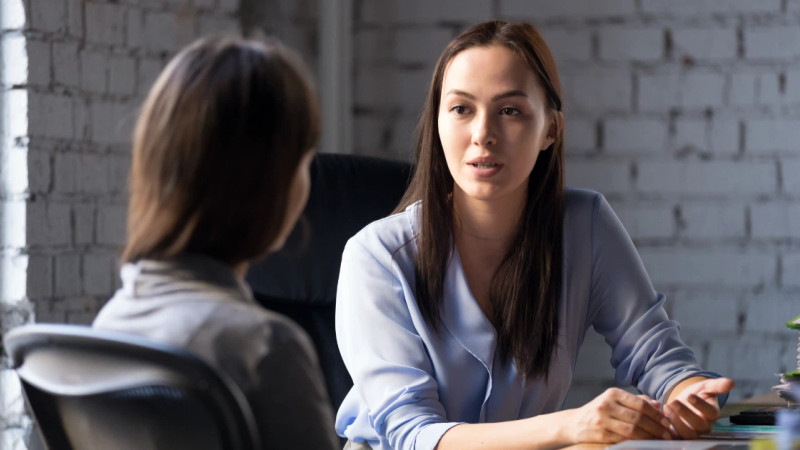 Two women in conversation across a desk in a whitewashed brick office, one facing camera in a light blue shirt