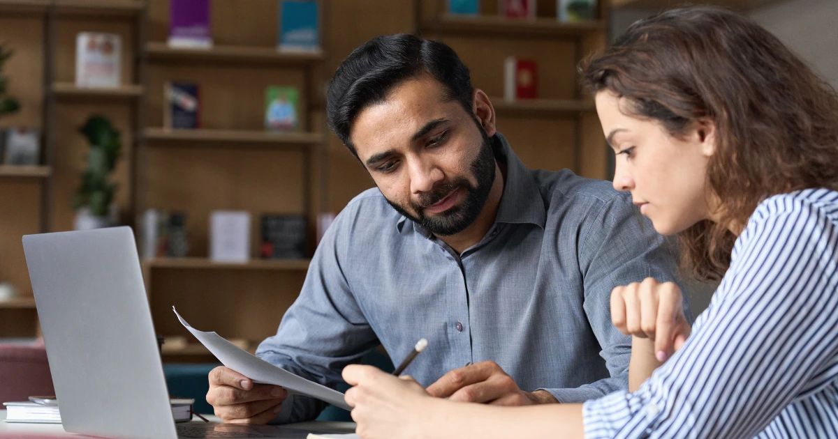 Man and woman reviewing a printed document together at a desk with a laptop and bookshelf behind them