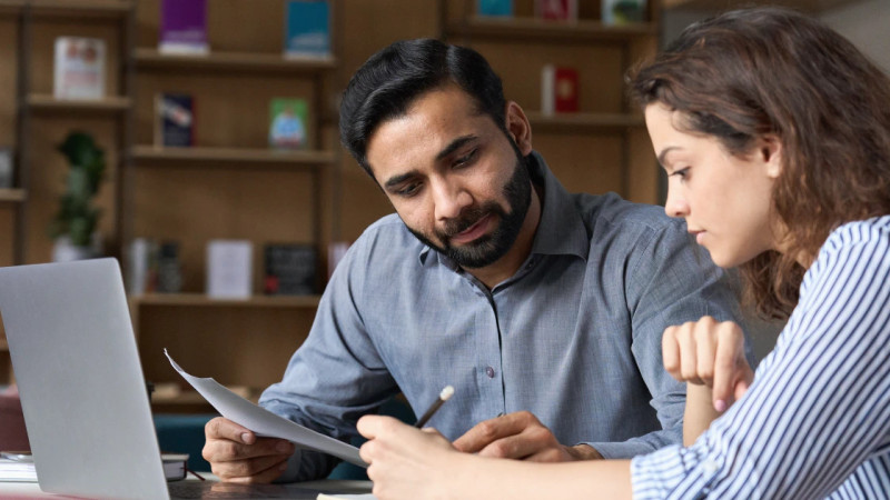Man and woman reviewing a printed document together at a desk with a laptop and bookshelf behind them