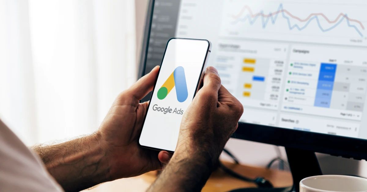 Hands holding a smartphone displaying the Google Ads logo, with a campaign dashboard visible on a monitor behind