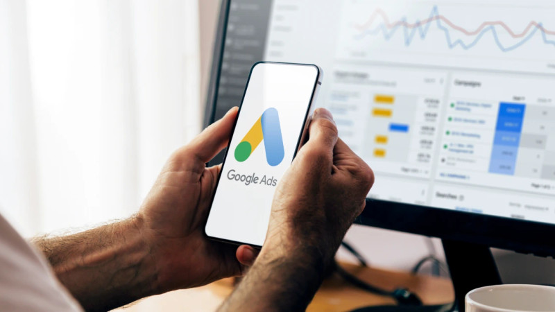 Hands holding a smartphone displaying the Google Ads logo, with a campaign dashboard visible on a monitor behind
