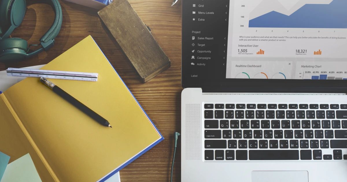 Overhead flatlay of a laptop showing a sales dashboard, yellow notebook, pencil, ruler and headphones on a wooden desk