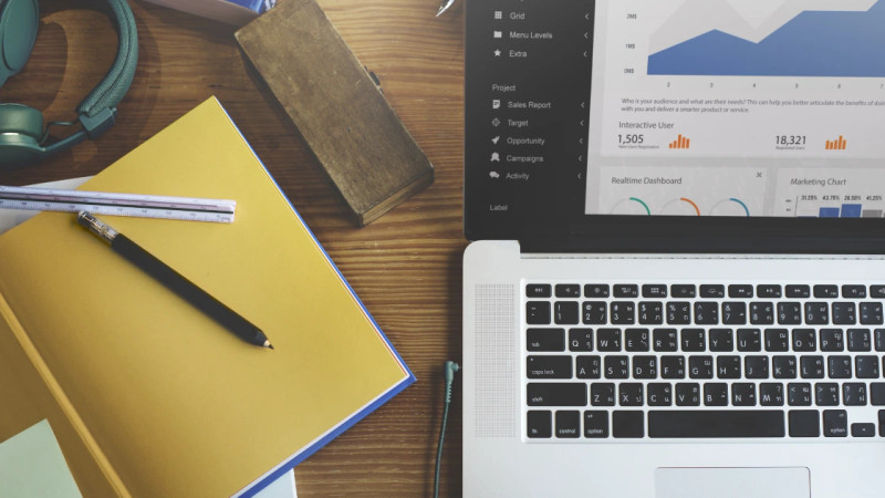 Overhead flatlay of a laptop showing a sales dashboard, yellow notebook, pencil, ruler and headphones on a wooden desk