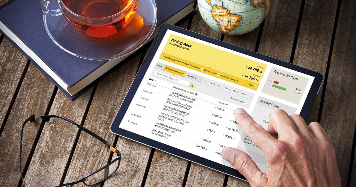 Hand scrolling through a bank account transaction list on a tablet, with tea, glasses and a globe on a wooden desk