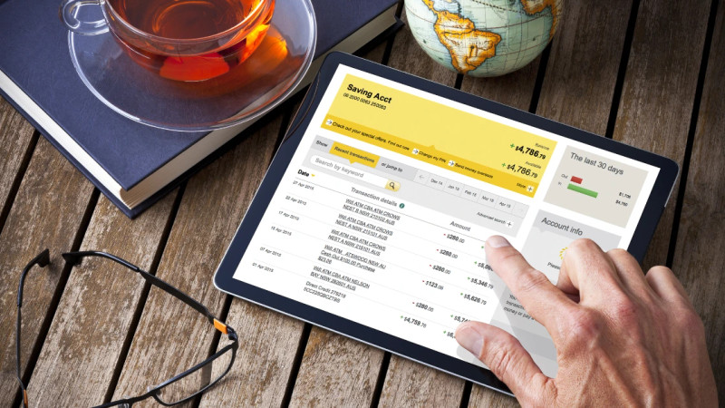 Hand scrolling through a bank account transaction list on a tablet, with tea, glasses and a globe on a wooden desk