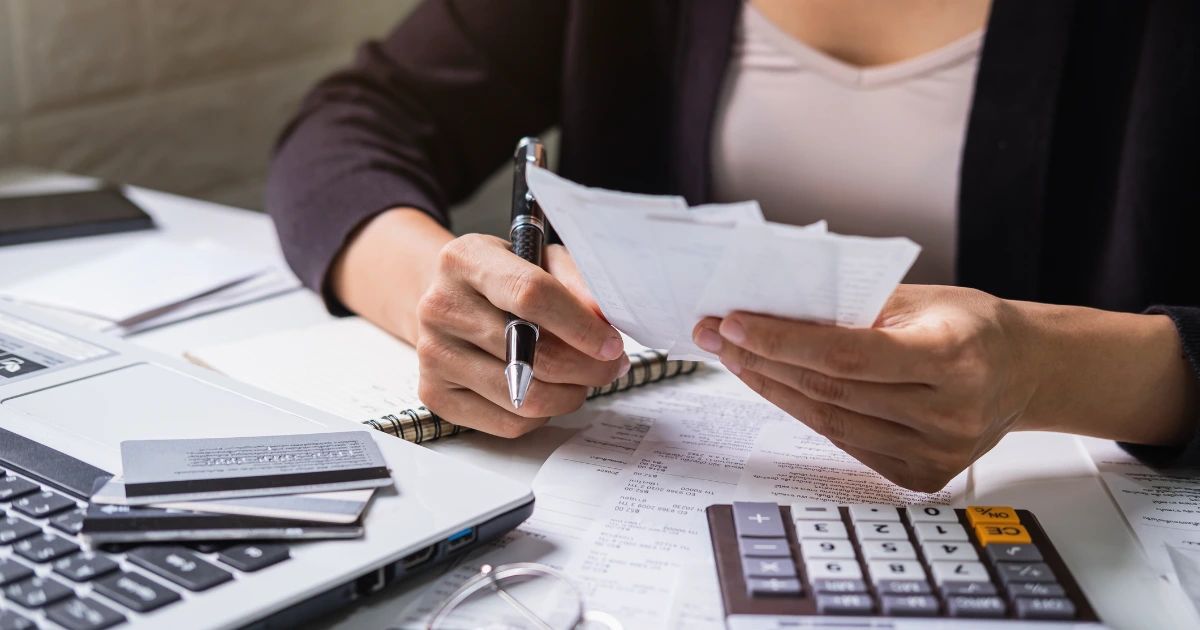 Woman holding receipts and pen over a desk with calculator, financial documents, credit cards and a laptop