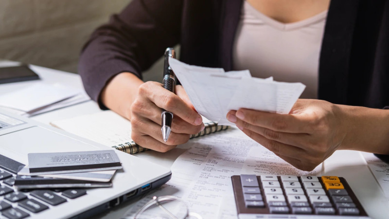 Woman holding receipts and pen over a desk with calculator, financial documents, credit cards and a laptop