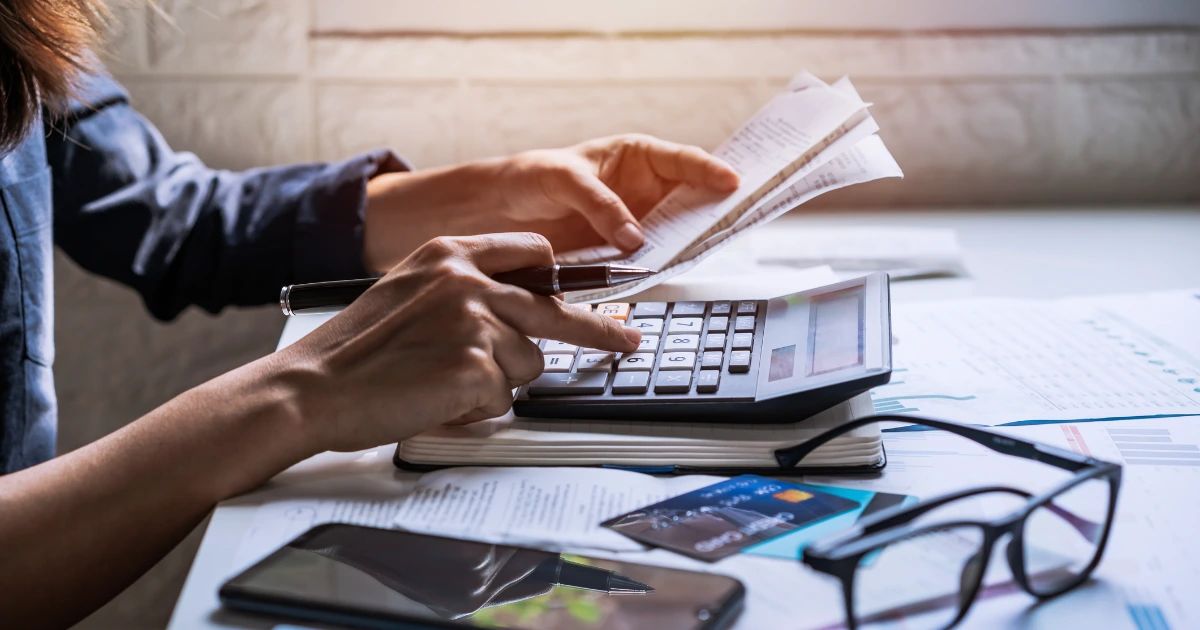 Person holding receipts and using a calculator at a desk with financial papers, credit cards and glasses nearby