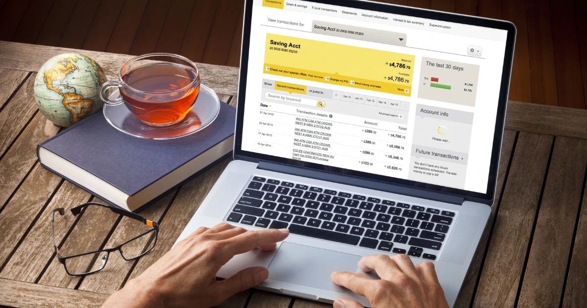Hands on laptop showing online banking savings account transactions, cup of tea and glasses on wooden desk