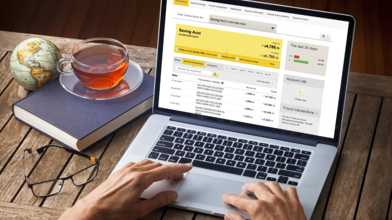 Hands on laptop showing online banking savings account transactions, cup of tea and glasses on wooden desk