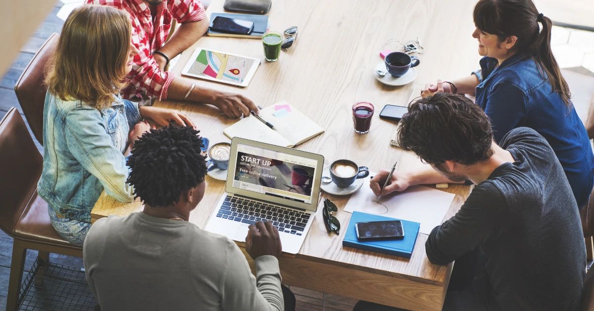 Four young professionals around a wooden table with laptop, tablet, notebooks and coffees in a casual café setting.