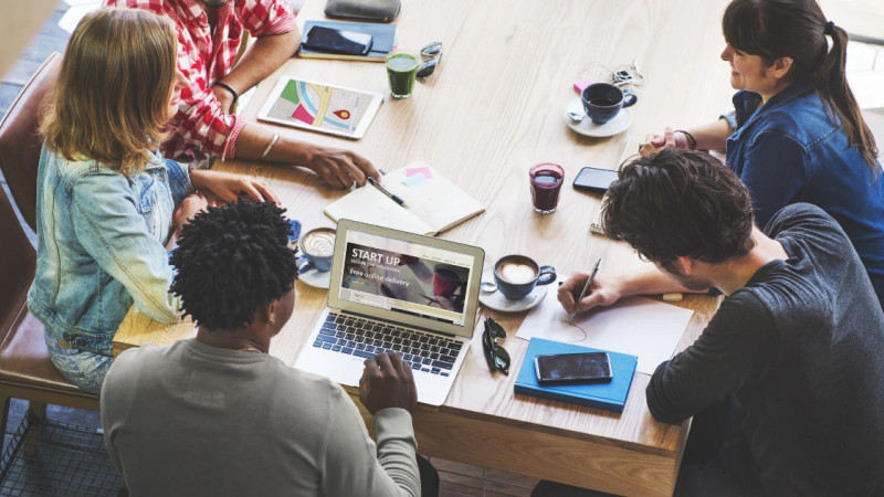 Four young professionals around a wooden table with laptop, tablet, notebooks and coffees in a casual café setting.